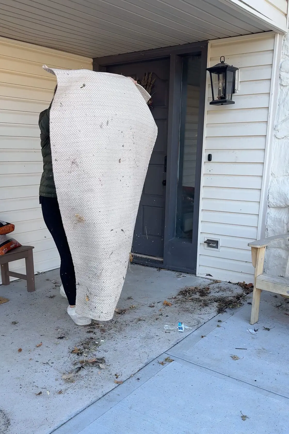 A person shaking out a large outdoor rug on a front porch, surrounded by scattered leaves and dirt, with a gray front door and white siding in the background.